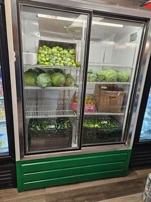 Two-door commercial display refrigerator with glass panels and green base, stocked with fresh produce on wire shelves