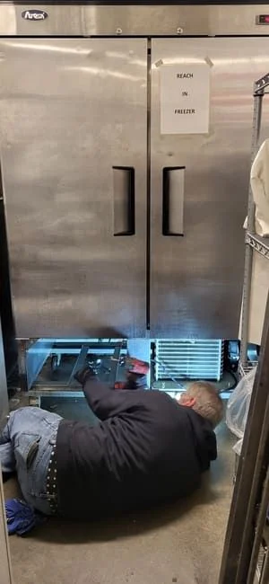 A commercial reach-in refrigerator with its access panel open being inspected by a technician inside a Cincinnati restaurant kitchen. The technician examines the compressor and condenser coil components during a diagnostic repair visit.