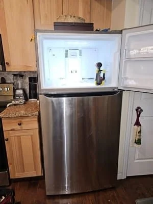 A residential refrigerator with its doors open showing organized, well-cooled interior contents in a Cincinnati home kitchen. An appliance repair technician inspects the lower condenser area during a refrigerator repair service call.